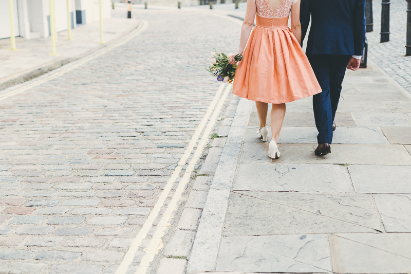 Bride and groom walking down the street in Old Portsmouth, form behind, bride wears peach, coral coloured wedding dress with cream beaded shoes and bouquet. Groom wears blue suit.
