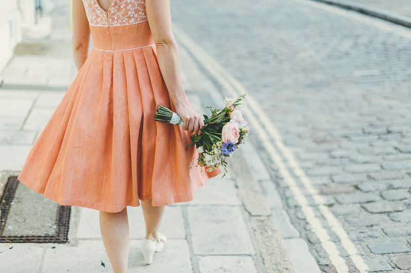 Bride and groom walking down the street in Old Portsmouth, bride wears peach, coral coloured wedding dress with cream beaded shoes and bouquet. Groom wears blue suit.