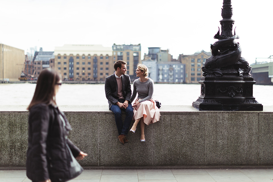 Couple sitting on the wall of the Thames in London, she wears pink satin skirt and grey jumper with colourful floral heels.
